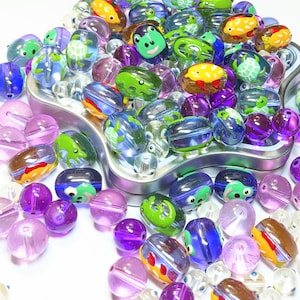 May include: A variety of colorful glass beads with different animal designs, including turtles, fish, and frogs. The beads are in a metal container and some are scattered on a white surface.