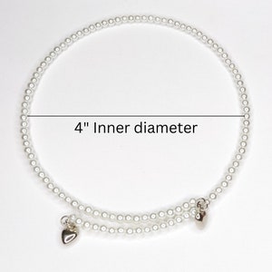 K&ouml;nnte beinhalten: Ein wei&szlig;es Perlenarmband mit zwei silbernen Herzanh&auml;ngern. Das Armband hat einen Innendurchmesser von 10 cm.