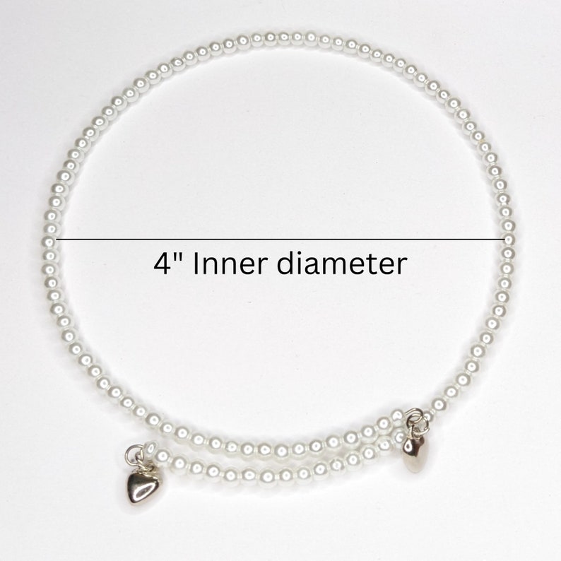 K&ouml;nnte beinhalten: Ein wei&szlig;es Perlenarmband mit zwei silbernen Herzanh&auml;ngern. Das Armband hat einen Innendurchmesser von 10 cm.