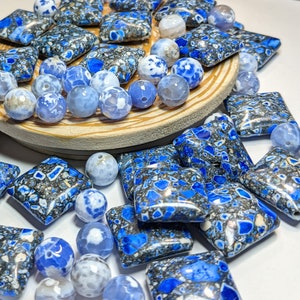 May include: A collection of blue and white agate gemstone beads with a marbled pattern. The beads are arranged in a circle on a wooden surface.