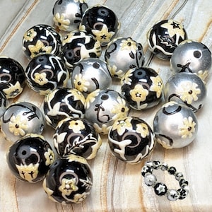 May include: A collection of black and silver beads with white floral designs. The beads are arranged in a random pattern on a wooden surface. There is also a silver bracelet with black and white floral designs.