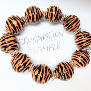 May include: A bracelet made of orange and black beads with a zebra print pattern. The beads are connected with silver spacers. The bracelet is on a white background with the text "Inspiration Sample" written in black.