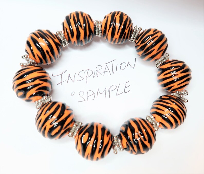 May include: A bracelet made of orange and black beads with a zebra print pattern. The beads are connected with silver spacers. The bracelet is on a white background with the text "Inspiration Sample" written in black.