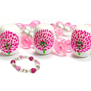 May include: A pink and white flower bead bracelet with a pink and white flower charm. The bracelet is made of white beads with pink flowers painted on them. The charm is a larger white bead with a pink flower painted on it.