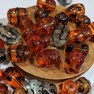 May include: A collection of orange and brown glass beads with black and white patterns. The beads are oval shaped and have a variety of designs, including stripes, dots, and swirls.