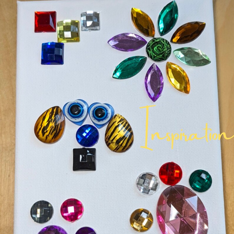 Puede incluir: Un lienzo blanco con brillantes y piedras preciosas de vidrio de colores dispuestos en un dise&ntilde;o caprichoso. El dise&ntilde;o incluye una flor, una cara de animal de dibujos animados y un grupo de piedras preciosas. La palabra "Inspiration" est&aacute; escrita en amarillo en el lienzo.