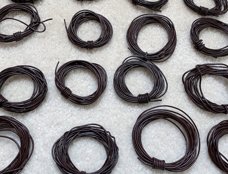 May include: A collection of 13 brown leather cords, each wound into a circle, arranged on a white surface.