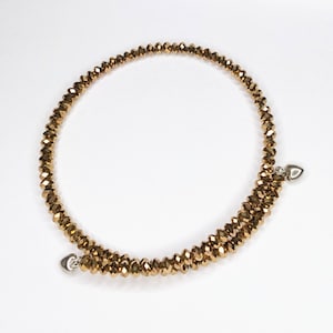 K&ouml;nnte beinhalten: Ein goldfarbenes Perlenarmband mit zwei silbernen Herzanh&auml;ngern. Das Armband besteht aus kleinen, facettierten Perlen und ist um sich selbst gewickelt.