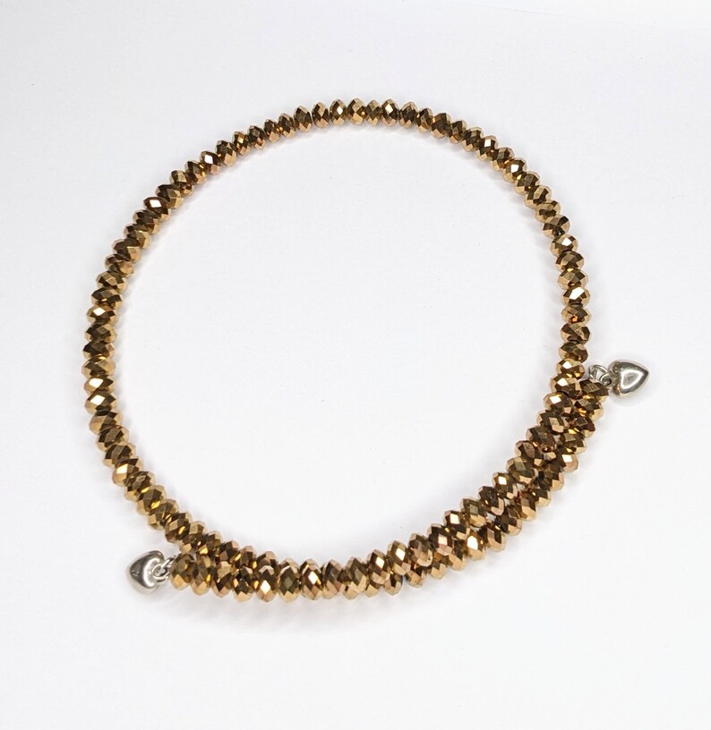 K&ouml;nnte beinhalten: Ein goldfarbenes Perlenarmband mit zwei silbernen Herzanh&auml;ngern. Das Armband besteht aus kleinen, facettierten Perlen und ist um sich selbst gewickelt.