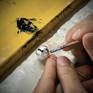 May include: A person is painting black dots on a white pearl with a small brush. The pearl is on a toothpick and the person is holding the toothpick with their left hand. The person is using their right hand to hold the brush.