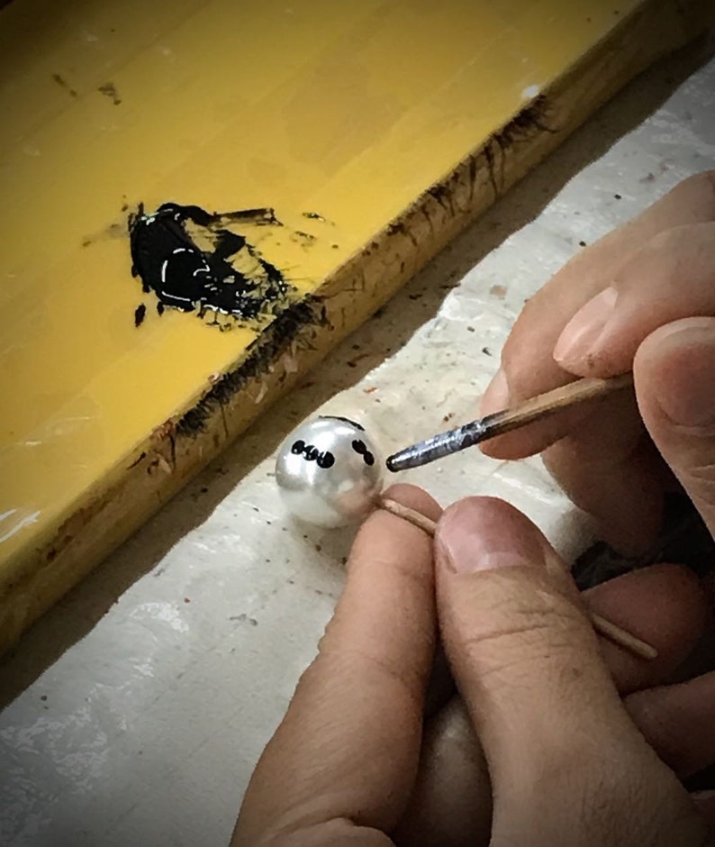 May include: A person is painting black dots on a white pearl with a small brush. The pearl is on a toothpick and the person is holding the toothpick with their left hand. The person is using their right hand to hold the brush.