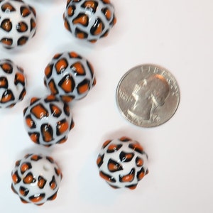 May include: Orange and white oval-shaped glass beads with a leopard print design. The beads are about the size of a UK 20 pence coin.