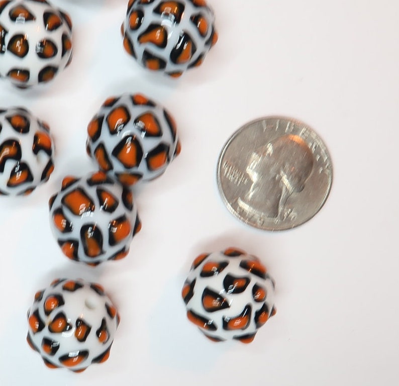 May include: Orange and white oval-shaped glass beads with a leopard print design. The beads are about the size of a UK 20 pence coin.