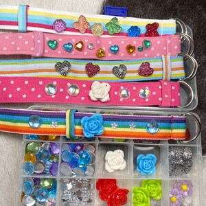 Puede incluir: Un conjunto de cinturones de tela coloridos con varios adornos, incluidos corazones, flores y pedrer&iacute;a. Los cinturones se muestran con un recipiente de pl&aacute;stico transparente lleno de pedrer&iacute;a, flores y otros adornos surtidos.
