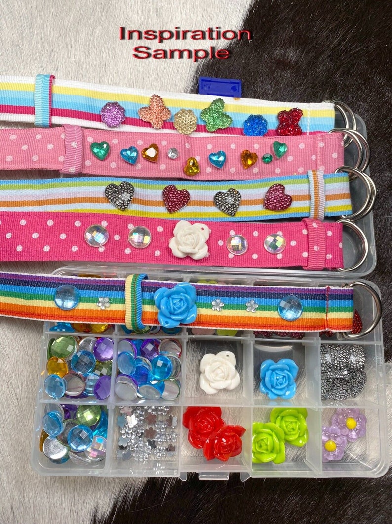 Puede incluir: Un conjunto de cinturones de tela coloridos con varios adornos, incluidos corazones, flores y pedrer&iacute;a. Los cinturones se muestran con un recipiente de pl&aacute;stico transparente lleno de pedrer&iacute;a, flores y otros adornos surtidos.
