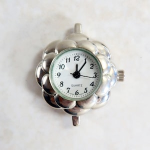 May include: A silver quartz watch pendant with a white face and black hands. The watch is surrounded by a silver, flower-like design. The watch face has black numbers and the word "QUARTZ" printed on it. The watch measures approximately 1 inch in diameter.
