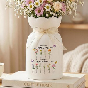 Op de afbeelding: Een witte keramische vaas met een geschulpte rand, vastgebonden met een beige lint, gevuld met kleurrijke bloemen. De vaas is versierd met de woorden "First Mom - Now Grandma" en bloemenillustraties.