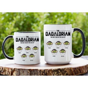 Op de afbeelding: Twee witte keramische mokken met zwarte handvatten en binnenkanten, elk voorzien van de tekst THE DADALORIAN THE BEST DAD IN THE GALAXY. Onder de tekst staan vijf illustraties van groene wezens in pods, gelabeld met de namen Rose, Daisy, Oliver, Violet en Rowan.