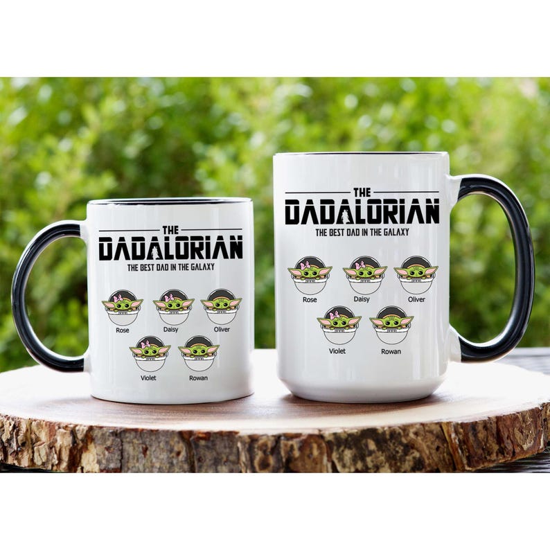 Op de afbeelding: Twee witte keramische mokken met zwarte handvatten en binnenkanten, elk voorzien van de tekst THE DADALORIAN THE BEST DAD IN THE GALAXY. Onder de tekst staan vijf illustraties van groene wezens in pods, gelabeld met de namen Rose, Daisy, Oliver, Violet en Rowan.