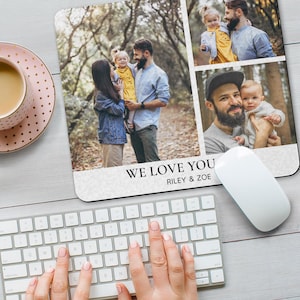 Può includere: Tappetino per mouse personalizzato con un collage di foto di famiglia. Il tappetino ha il testo "WE LOVE YOU" e i nomi "RILEY & ZOE" stampati sopra. Un mouse e una tastiera per computer bianchi sono in primo piano, con una tazza da tè rosa e dorata nell'angolo in alto a sinistra.