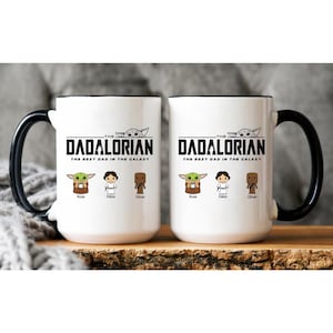 May include: Two white ceramic mugs with black handles and rims. The design features the text THE DADALORIAN THE BEST DAD IN THE GALAXY with a small character graphic above. Below are three cartoon figures labeled Rose, Daisy, and Oliver.