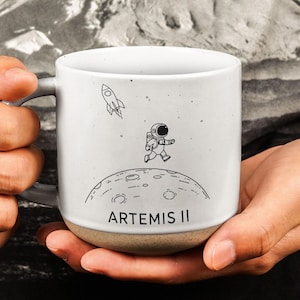 Puede incluir: Taza de cerámica blanca con asa gris y base beige. La taza presenta una ilustración negra de un astronauta corriendo sobre una luna, un cohete y el texto "ARTEMIS II".