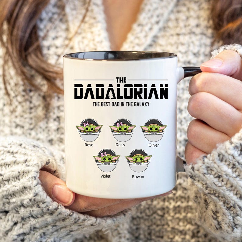 Op de afbeelding: Een witte keramische mok met een zwarte binnenkant en handvat. De voorzijde bevat de tekst THE DADALORIAN THE BEST DAD IN THE GALAXY boven vijf illustraties van groene wezens in pods, gelabeld met de namen Rose, Daisy, Oliver, Violet en Rowan.