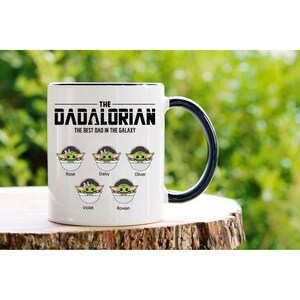 Op de afbeelding: Een witte keramische mok met een zwarte handgreep en rand, voorzien van de tekst THE DADALORIAN THE BEST DAD IN THE GALAXY. Onder de tekst staan vijf illustraties van groene wezens in pods, elk gelabeld met de namen Rose, Daisy, Oliver, Violet en Rowan.