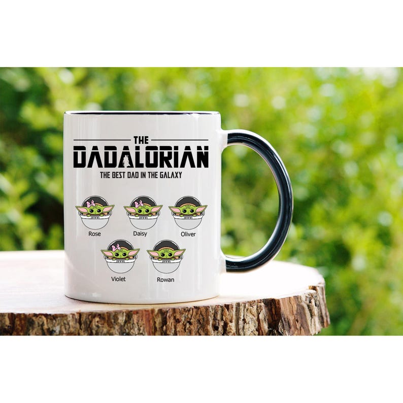 Op de afbeelding: Een witte keramische mok met een zwarte handgreep en rand, voorzien van de tekst THE DADALORIAN THE BEST DAD IN THE GALAXY. Onder de tekst staan vijf illustraties van groene wezens in pods, elk gelabeld met de namen Rose, Daisy, Oliver, Violet en Rowan.