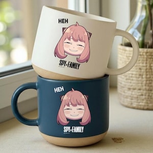 Puede incluir: Dos tazas de cerámica apiladas. La taza superior es blanca y la inferior es azul oscuro. Cada taza presenta una ilustración de un personaje de pelo rosa, con el texto "HEH" y "SPY x FAMILY".