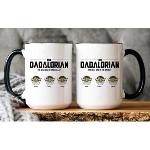 Op de afbeelding: Twee witte keramische mokken met zwarte handvatten en randen. De voorzijde bevat de tekst "THE DADALORIAN THE BEST DAD IN THE GALAXY" boven drie illustraties van groene wezens in pods, gelabeld met de namen "Rose", "Daisy" en "Oliver".