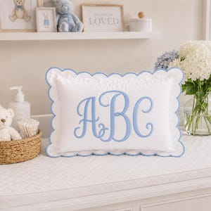 May include: A rectangular white pillow featuring a scalloped edge trimmed in light blue thread. The center of the pillow displays the embroidered letters ABC in a large, flowing, light blue script font, centered on the white fabric surface.