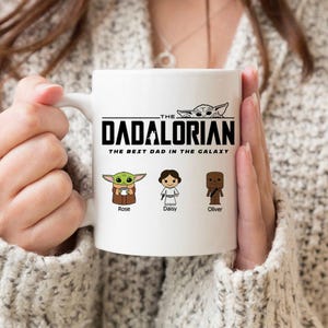 May include: A white ceramic mug featuring the black text THE DADALORIAN THE BEST DAD IN THE GALAXY. Below the text are three cartoon characters with the names Rose, Daisy, and Oliver printed underneath each respective figure in black ink.