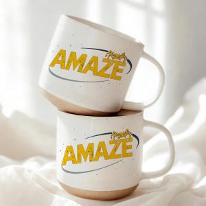 Puede incluir: Dos tazas de cerámica blanca apiladas. Cada taza tiene una base beige y una sección superior blanca. La palabra "AMAZE" está impresa en amarillo, con un diseño de cangrejo. Una línea curva gris subraya la palabra.