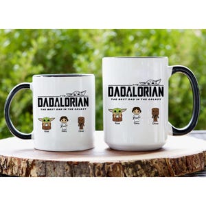 May include: Two white ceramic mugs with black handles and rims. The text reads THE DADALORIAN THE BEST DAD IN THE GALAXY. Below the text are three cartoon characters labeled Rose, Daisy, and Oliver, with a small creature graphic above the text.