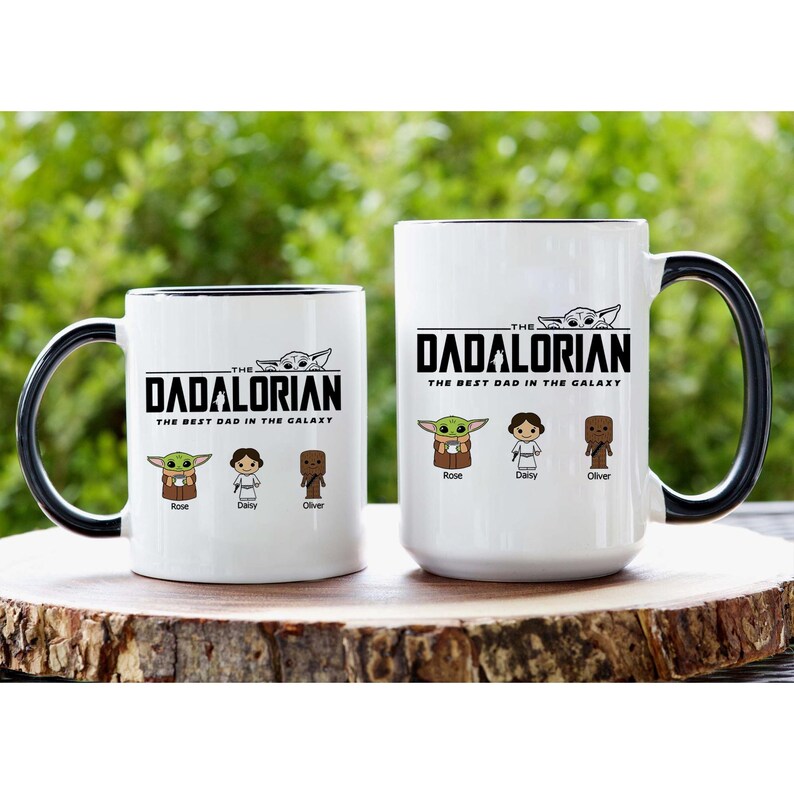 May include: Two white ceramic mugs with black handles and rims. The text reads THE DADALORIAN THE BEST DAD IN THE GALAXY. Below the text are three cartoon characters labeled Rose, Daisy, and Oliver, with a small creature graphic above the text.