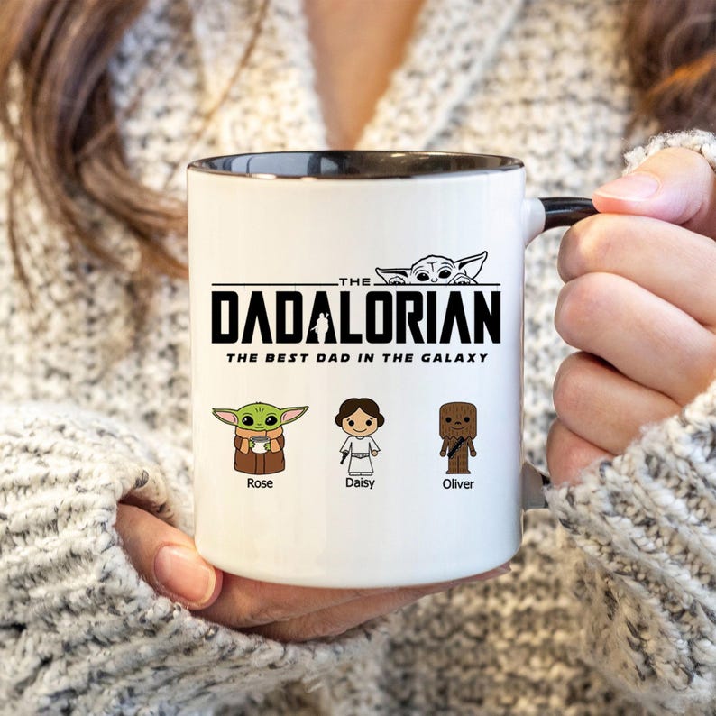May include: A white ceramic mug with a black interior and handle. The front features the black text THE DADALORIAN THE BEST DAD IN THE GALAXY with a small character graphic above. Below are three cartoon figures labeled Rose, Daisy, and Oliver.