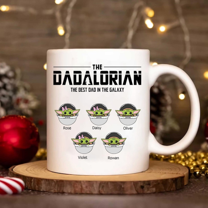 Op de afbeelding: Een witte keramische mok met de zwarte tekst THE DADALORIAN THE BEST DAD IN THE GALAXY boven vijf illustraties van groene wezens in pods, gelabeld met de namen Rose, Daisy, Oliver, Violet en Rowen in een zwart lettertype.