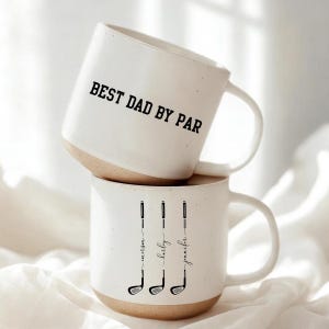 May include: Two white ceramic mugs with a tan base. The top mug reads "BEST DAD BY PAR" in black text. The bottom mug features three golf club illustrations with names below them.
