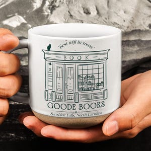 Puede incluir: Taza Goode Books de cerámica blanca con asa gris y base beige. La taza presenta una ilustración verde de una librería con el texto "best wifi in town" y "GOODE BOOKS Sunshine Falls, North Carolina."