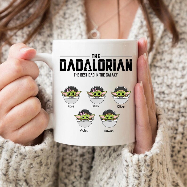 Op de afbeelding: Een witte keramische mok met de zwarte tekst THE DADALORIAN THE BEST DAD IN THE GALAXY. Onder de tekst staan vijf illustraties van groene wezens in pods, gelabeld met de namen Rose, Daisy, Oliver, Violet en Rowan in een zwart lettertype.