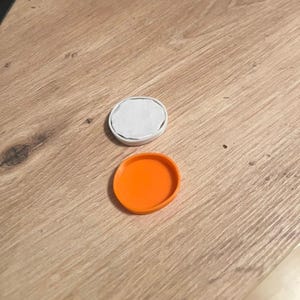Könnte beinhalten: Zwei kleine, runde Objekte liegen auf einer Holzoberfläche. Eines ist eine leuchtend orangefarbene Scheibe, das andere ein weißes, kreisförmiges Objekt mit grauem Innenraum. Die Artikel scheinen Deckel oder Behälter zu sein, möglicherweise für kleine Gegenstände oder zur Aufbewahrung.