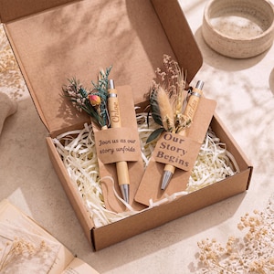 May include: Two light wood pens with silver accents, each attached to a kraft paper card inside a cardboard box filled with paper shreds. One card reads Join us as our story unfolds, the other Our Story Begins. Dried flowers adorn each pen.