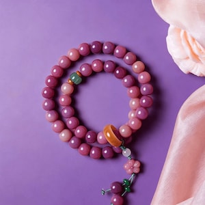 May include: A strand of purple and pink beaded jewelry with a flower and bead pendant. The beads are round and vary in shade. An orange ring and green bead accent the design. The background is a solid purple.