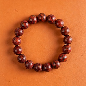 May include: A circular bracelet composed of smooth, spherical beads. Each bead features a mottled pattern of deep red, dark brown, and subtle black tones, creating a variegated appearance across the entire piece of jewellery.