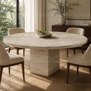 Puede incluir: Una mesa de comedor redonda de travertino beige con una base de pedestal central gruesa, acompañada de cuatro sillas con asientos tapizados en color crema y marcos de madera oscura, dispuestas en una habitación luminosa.