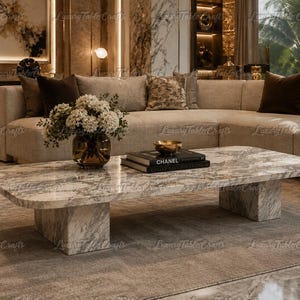 May include: A rectangular coffee table with rounded corners, crafted from white and grey veined marble. The table features two thick, block-style pedestal legs and supports a glass vase filled with white flowers and a stack of two black books.