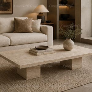 Op de afbeelding: Een rechthoekige beige salontafel met een steenachtig oppervlak, ondersteund door twee rechthoekige sokkels. De tafel bevat boeken, een kom en een vaas met groen. Een beige bank en vloerkleed maken de setting compleet.