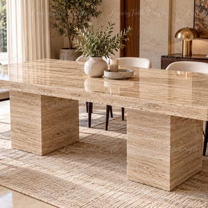 Pode incluir: Uma mesa de jantar retangular feita de pedra travertino bege polida, apresentando uma superfície lisa e brilhante com veios naturais e duas bases de pedestal robustas em forma de bloco que sustentam o amplo tampo da mesa.