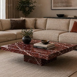 May include: A square coffee table made of deep red marble with prominent white veining. The table features a thick, flat top supported by a central block base. On the surface sits a small black vase with greenery and two stacked books.